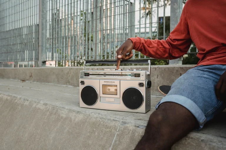 Crop anonymous black guy in denim shorts sitting on concrete step near metal railing and pressing button on old fashioned cassette recorder — an editorial image for "Contemporary R&B and the Sound of the 1990s."