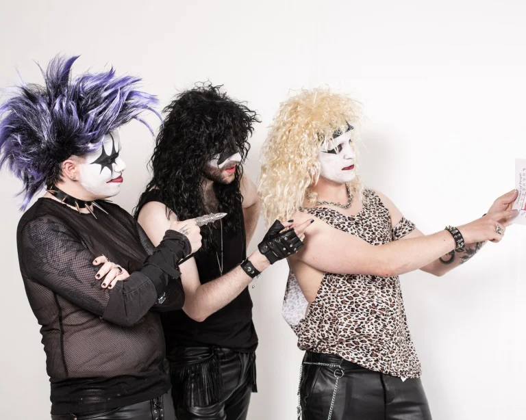 Three band members dressed in glam rock style with dramatic face paint together indoors — an editorial image for "David Bowie and the Art of Musical Reinvention."
