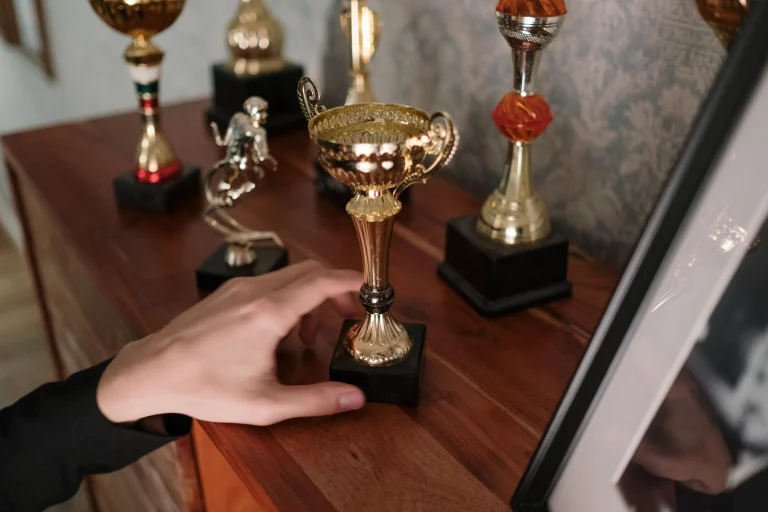 A close-up of a hand reaching for a shiny trophy on a wooden shelf, symbolizing success — an editorial image for "The Grammy Awards and the Shaping of Musical Prestige."