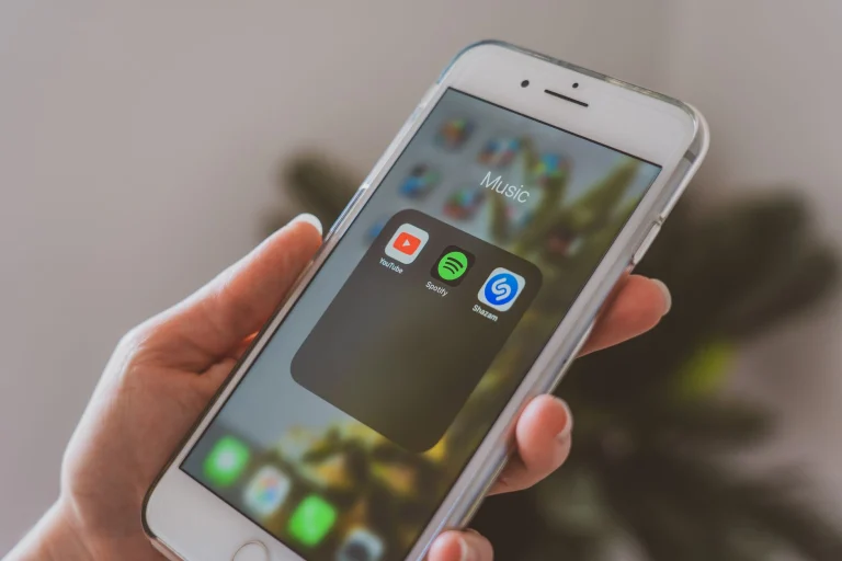 Hand holding a smartphone displaying popular music apps like Spotify and Shazam in a close-up shot — an editorial image for "How Streaming Reshaped the Music Business."