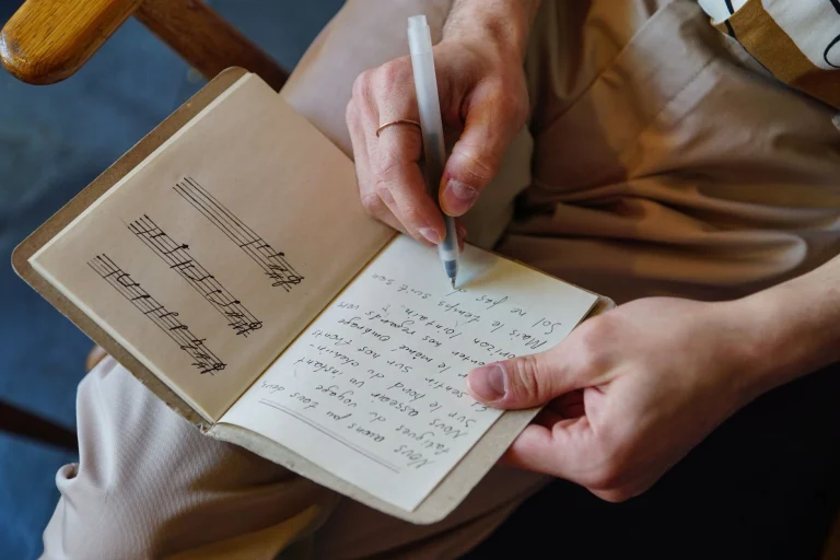 Male songwriter writes lyrics and music notes in a notebook, capturing the creative process — an editorial image for "I Will Always Love You and the Power Ballad."