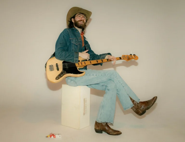 Man dressed in cowboy hat and denim, holding a bass guitar, sitting on a studio box — an editorial image for "Rumours: How Fleetwood Mac Created a Landmark Album."