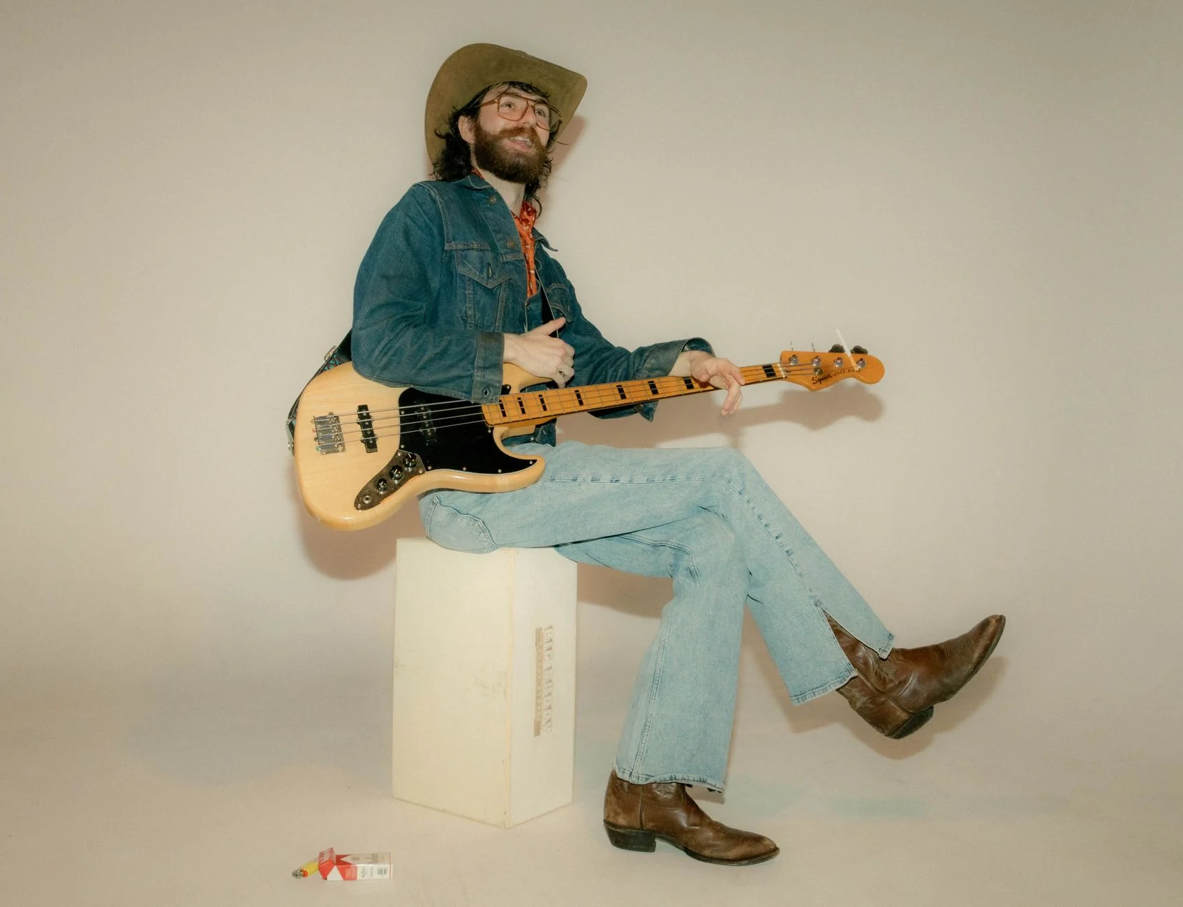 Man dressed in cowboy hat and denim, holding a bass guitar, sitting on a studio box — an editorial image for "Rumours: How Fleetwood Mac Created a Landmark Album."