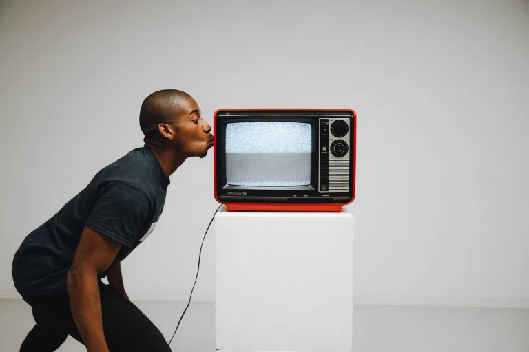 A man playfully kisses a vintage red television in a minimalist studio setting, evoking nostalgia and retro vibes — an editorial image for "Whitney Houston: Vocal Power and Crossover Stardom."